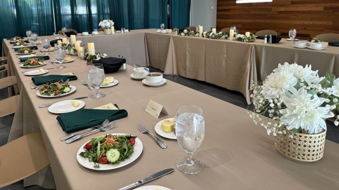 Long table draped in beige tablecloth with formal dinner setup.