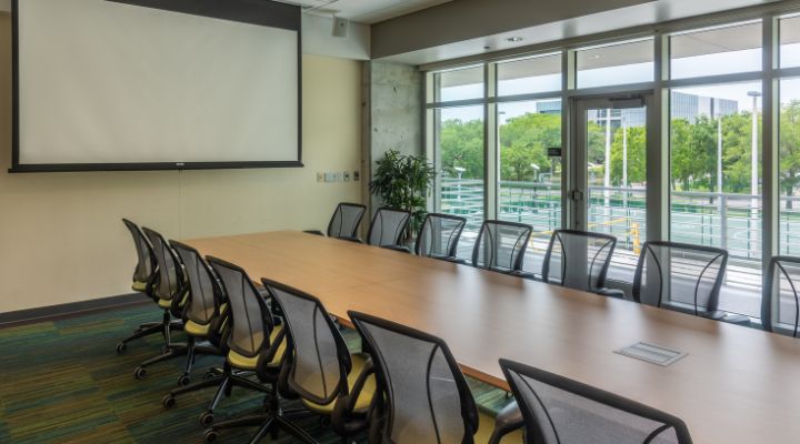 A long conference table surrounded by chairs with a projector hanging to the left