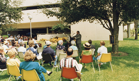 People in chairs on the lawn while a person speaks at a lecturn