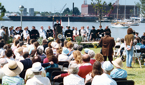 People sitting in chairs along while a person in the background gives a speech in front of a podium in front of Bayboro Harbor