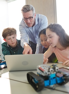 Three students sitting at a table looking at a laptop with a robotic vehicle in front and a teacher looking over them from behind