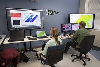 Two students sitting in front of a table that has multiple laptops and computer screens