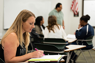 A student sitting at a desk looking at an open notebook with a group of students and an instructor in the background