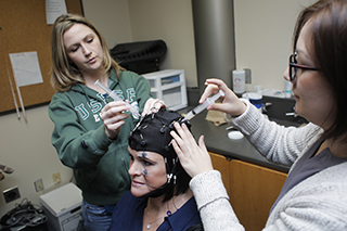 A person sitting wearing an experimental cap with two people on either side holding holding syringes up to the cap