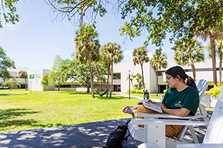 female student reading a book sitting in a chair outside