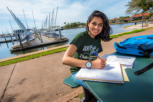 A female USF student sitting by the water and taking notes.