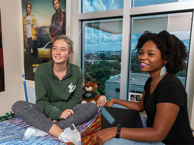 Two female USF students sitting on a bed in a dorm room.