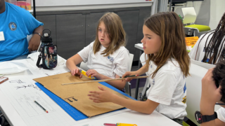 Two students listening and working on a project, one holding a ruler