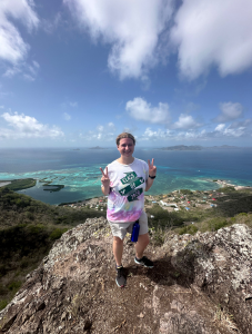Student holding up double peace signs while on a high rock with blue water behind her.
