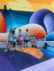 Students standing and playing in front of colorful mural wearing "USF Camp Rocky" tee shirts