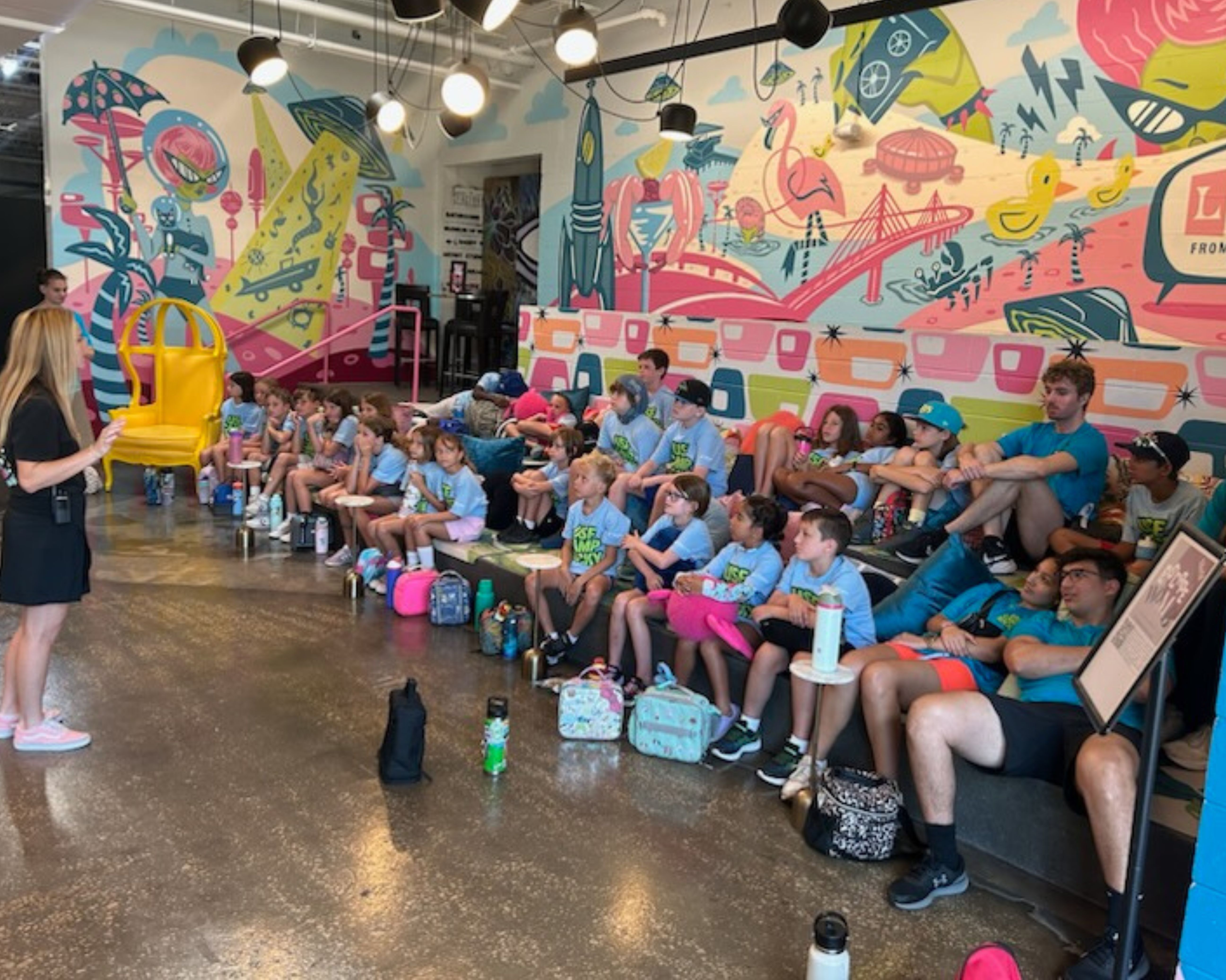 Group of students listening to a speaker while sitting on bleacher seats in front of a mural