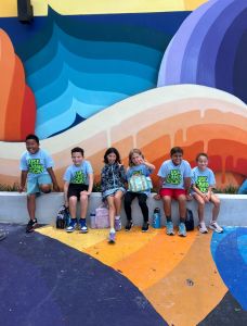 Students standing and playing in front of colorful mural wearing "USF Camp Rocky" tee shirts