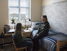 Student sitting on a bed talking to a student sitting in a chair