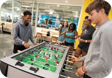 Two students playing foosball with other students looking on