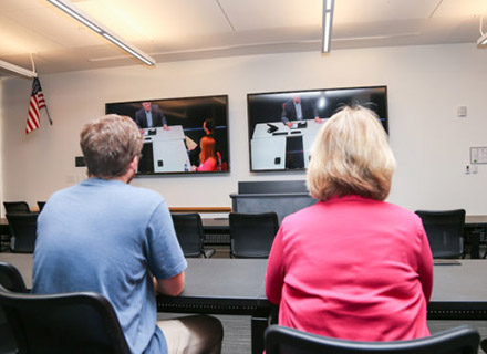 Two people with their back to the camera watching two high definition monitors in a classroom.