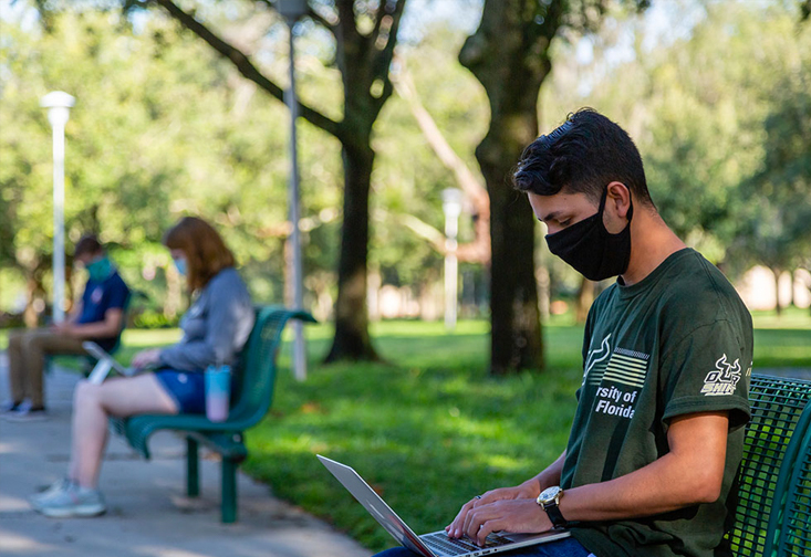 Students wearing masks while sitting on benches while working on their laptops
