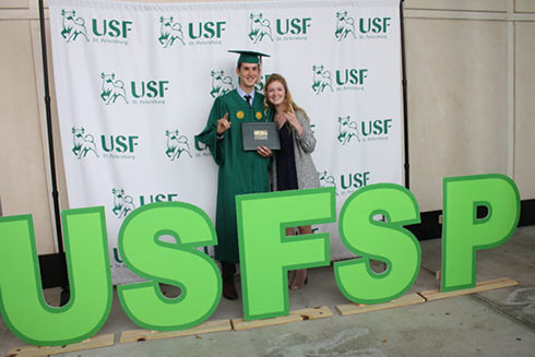 Student in graduation cap and gown posing with mother in front of USFSP sign