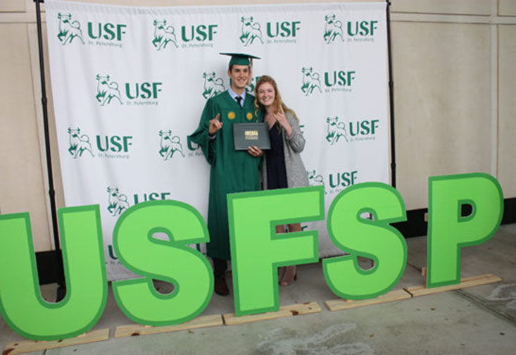 Student in graduation cap and gown posing with mother in front of USFSP sign