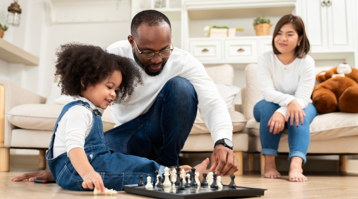 A man and a small child playing chess on the floor while a woman watches and smiles while sitting on the couch