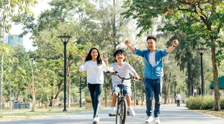 Two parents and child outside. This child is riding a bike with encouragement from parents