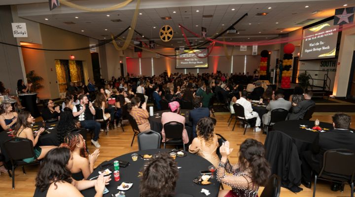 Students in large ballroom sitting at tables wearing formal clothes. 